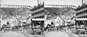 « Près de ville et terrace » - On y voit un marché de la basse-ville, les murs de la citadelle en arrière-plan, les bâtiments commerciaux de J. Hinds, d'A.W. Lebel et de James A. Quinn ainsi qu'un stand de légumes en avant-plan ca. 1860's
