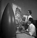Laboratoires du Conseil de recherches pour la défense. John Mar (assis) et Gordon Philips avec la coiffe qui contiendra les instruments pour mettre à l'essai le déploiement ingénieux de l'antenne de 75 pieds du satellite canadien « TopsideSounder » March 1961.
