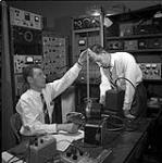 Defence Research Board Laboratories. Engineer R.J. Bibby (standing) and technician Roy Cunningham with mock-up of ground station readied to receive information from "Topside Sounder" satellite Mar. 1961