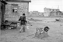 Unidentified Cree carrying pails on yoke walking past building 1973