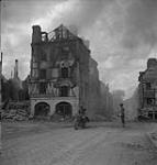 Unidentified member of the Canadian Provost Corps directing traffic past a burning building 17 Aug. 1944