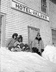 Jimmy Komooak, chief herder of reindeer, standing under the Hotel Aklavik sign, with two children March 1956.
