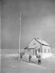 Tapatai and Reverend W.J.R. James raising the White Ensign outside the Anglican Rectory, marking the presence of Canadian Army personnel taking part in Exercise Muskox, Baker Lake (Qamanittuaq), Northwest Territories [Nunavut], March 1946 March 1946.