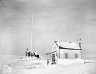Rev. W.J.R. James (right) and his Inuit assistant, Tapatai (left), outside St. Aidan's Anglican Mission and Rectory, Baker Lake, Northwest Territories, [(Qamanittuaq), Nunavut], March 1946 March 1946.