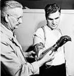 Private Ed Gerris of the Royal Canadian Army Service Corps, seen here with Principal Eccles of Shaw Business Schools, is using a specially-designed glove-covered artificial hand to learn penmanship during a retraining course on Primary Accountancy 1944