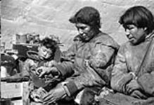 Inuit family inside an Igloo. The woman [Ikualaaq] is cutting meat with an ulu 1948-1953.