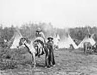 [Two unidentified Cree wearing blankets and posing in front of teepees. One is on a horse.]. Original title: Cree Indians [between 1870-1910]