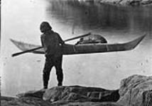 Inuit man carrying his kayak to the water to start on hunt. [Greenland] 1872-1873.