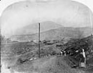 On the road of Lytton - woman with a bucket and a washboard and two men horseback riding. View of telegraph posts 1865