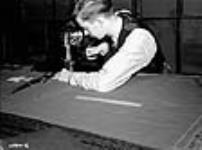 Un homme guide une coupeuse de toile automatique sur une pile de tissu kaki le long d'un patron d'uniforme canadien tracé à la craie Dec. 1939