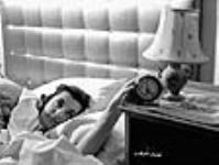 Woman munitions worker (prob. for Robert Mitchell Co.) in bed reaches to turn off alarm clock Mar. 1941