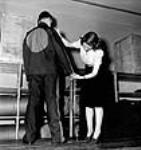 Woman worker takes measurement of the sleeve of a man modelling internment uniform at the Standard Overalls Company Mar. 1943