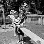 Deux enfants, dont la mère, Mme Jack Wright, travaille dans un poste à temps partiel, jouent sur une balançoire à bascule avec une jardinière d'enfants Sept. 1943
