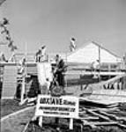 Vue d'ouvriers construisant une maison préfabriquée pour les travailleursde guerre M. et Mme Harry Kalek derrière une pancarte affichant « Loxtave Homes » sur laquelle une horloge indique 14 heures 50 mai 1944