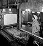 L'ouvrière Marie Geoffrion travaillant sur la machine à découper la cordite derrière une plaque de verre armé aux usines Cherrier et Bouchard de la fabrique de munitions Defense Industries Limited juil. 1944