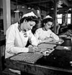 Female workers Vera Mann and Marie Grondin shown discing detonator pellets, making the seal airtight on missiles in the Cherrier and Bouchard plants of the Defense Industries Limited July 1944