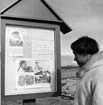 Unidentified Inuit man looking at a National Film Board release posted on the outdoor notice board in Cape Dorset August 1961.