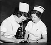Miss Holmes, nurse in training, looking into microscope while Miss S. Wray, R.N., watches at St. Joseph's General Hospital in Port Arthur, Ont c 1955