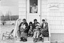 Mrs. Pulak, nurse at the Lansdowne nursing station, is watching from inside while [Indigenous] women and children sit on the front steps of the station June 1957