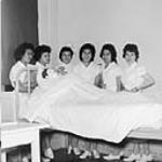 [Indigenous] nurses-in-training [unknown, Carole Maitland (nee Daniels), Doris Mitchell-Smith, Mae Wilson, Frances Wolfgang (nee Stewart), Delores Barton (nee Stewart)] are standing beside a bed with a dummy propped up on the pillows Nov. 1960