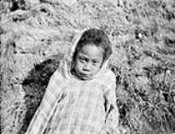 [Portrait of an unidentified child wearing a hooded shirt in front of a mound of dirt and grass, possibly Nunavut]. Original title : Half breed eskimo children [Negro], [Nunavut?] n.d.