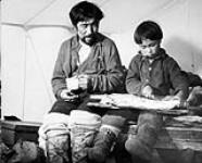 Inuk man watches boy cut salmon inside their tent at Chesterfield Inlet (Igluligaarjuk) 1946 or 1947.