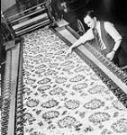 Man in a tie reaching over a bolt of fabric that is being produced by textile machinery 24 Feb. 1958
