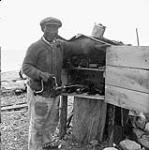 George Weetaltuk standing beside his outdoor tool cupboard. 1949.
