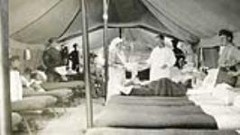 A nursing sister is assisting a doctor in a ward at the tent hospital. She is holding out a tray for him to take medical tools from 1916