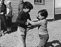 Two young Inuit boys play fighting while three Inuit women watch in the background, in Baker Lake (Qamanittuaq), Nunavut [the late David Annanaut is the boy on the right, the woman in the centre is the late Marjorie Esa and the woman on the right is Ruth Tulurialik] 1948.