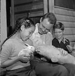 Leah Idlout, an Inuit girl from Pond Inlet, seated with Douglas Wilkinson and a boy outside of the Wilkinson's residence at Kingsmere, Quebec April, 1952.