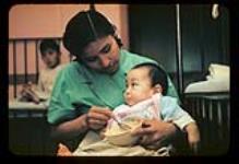 Nurse's aide feeding an infant at the Aklavik Anglican Hospital, Aklavik, N.W.T [ca. 1956]