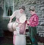 Woman dressed as a nineteenth century dairy farmer pours cream into a butter churn while a young boy watches as represented in the NFB production on the Canadian dairy industry August 1950
