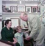 Elderly man and woman with little girl examining the contents of a jar, Kingsville November 1954