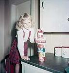 Little girl in kilt stands on chair and stacks canned goods from the Co-op on kitchen counter septembre 1953