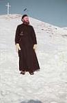Priest in a cassock at the bottom of a hill, a cross stands at the top of the hill, Northwest Territories 1957
