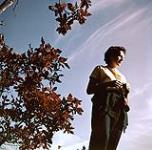 Portrait en contre-plongée d'une femme portant une chemise jaune. Branches d'un arbre à sa droite. Chalet Shilly Shally, parc de la Gatineau s.d.