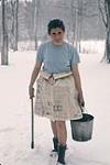 Helen Forsey standing in the snow wearing a skirt made of newspaper and holding a metal bucket in one hand. Shilly Shally Lodge, Gatineau Park 1965