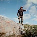 [Jimmy Spence, a Cree trapper and guide,] standing on a rocky slope and pointing, Norway House, Manitoba [between 1955 and 1963].