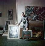 Robert Hubbard holding two paintings and standing in front of several paintings, at the National Gallery of Canada [entre 1955-1963]