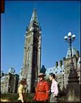 Man and woman talking with RCMP officer in front of Canadian Parliament buildings, Ottawa. [Un couple parle avec un agent de la GRC devant la les bureaux du parlement, Ottawa.] octobre 1949.