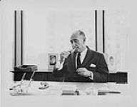 Man seated at a desk lighting a cigar [ca 1955-1976].