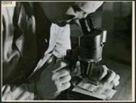 Un homme regarde dans un microscope en se servant d'un instrument spécialement conçu par les laboratoires de la GRC à Rockcliffe pour détecter les billets, l'écriture et les coupons d'essence contrefaits, à Rockcliffe June 1944