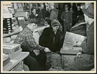 Dans le magasin général d'Harry Nedelle, deux clients conversent près du poêle à bois pendant que d'autres font la queue au comptoir à l'arrière-plan, à Fort-Coulonge n.d.