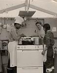 [Barbara Hinds at Cape Dorset Tourist Camp where a man and woman make doughnuts on a stove] [between 1956-1960]
