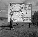 Et maintenant, où allons-nous? Audrey James debout à un grand panneau sur la route transcanadienne près de Kirkland Lake (Ontario) 1 août 1954.