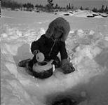 Boy using a kettle to collect water from a hole in a frozen lake, Brochet, Manitoba March, 1955.