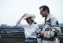 Cowgirl wearing white and man wearing knitted sweater holding a camera at the High River branding 1959