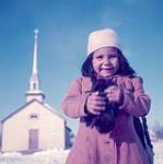 Jeune fille debout avec une église à l'arrière plan, Southend (Saskatchewan) [Rose Anne Hardotte (née Jobb) de la nation crie de Peter Ballantyne] mars 1955.