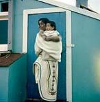 Woman wearing an amauti with her child in front of a wood framed building [Mary Ashoona with her daughter Pootoogoo Toonoo, Kinngait, Nunavut] [between June-September, 1960].